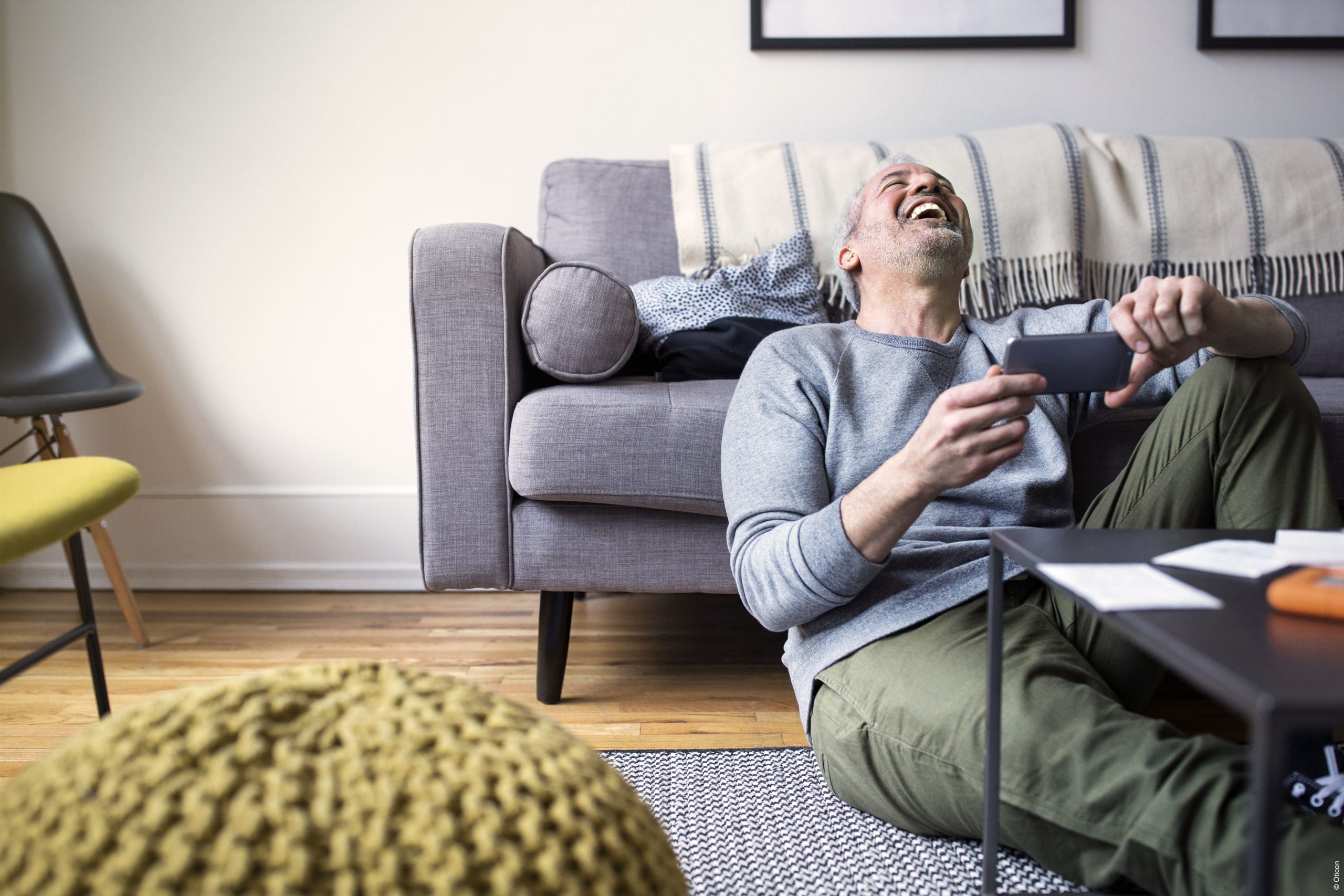 Mann hält Smartphone während er im Wohnzimmer vor seiner Couch sitzt und lacht